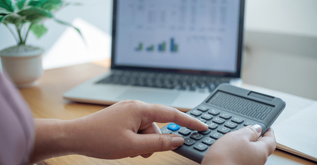 A pair of hands holding a calculator at a desk with a laptop open on a graph